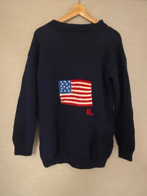 Mörkblå stickad tröja i RL-stil med USA-flagga  - En mörkblå stickad tröja med en detaljerad stickad amerikansk flagga på framsidan. Under flaggan finns bokstäverna 'RL' broderade. Tröjan har ribbstickade muddar och nederkant. Tröjan har stickats av en äkta farmor och tog ungefär 40 timmar plus 200 kronor i material att göra. Knappt använd.   American Flag.   Notera att detta INTE är en Ralph Lauren utan en hemmagjord tröja.   Möjligt att hämta upp fraktfritt i Östermalm eller Vallentuna.