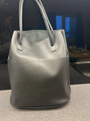 Svart tote handväska i läderimitation - Stilren svart tote handväska med två långa handtag och rymligt huvudfack. Väskan har minimalistisk design, stora ytterfickor och innerfack med dragkedja. Tillverkad i slät läderimitation och passar perfekt för dig som gillar enkel och modern stil.