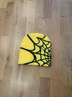 Gul mössa med spindelnätsmönster - Cool gul mössa med svart spindelnätsmönster. Mössan har en klassisk passform och är tillverkad i mjukt, stretchigt material som sitter skönt på huvudet. Perfekt för dig som vill sticka ut med en unik accessoar.