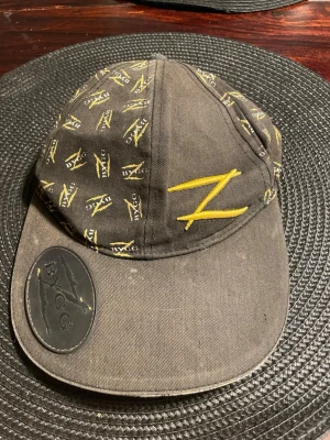 Svart BYGG keps med gul brodyr - Säljer en svart keps från BYGG med gul broderad Z-logga framtill och små mönstrade Z-detaljer runtom. Kepsen har en rundad skärm och ett stort BYGG-märke i gummi på skärmen. Materialet är bomullstyg och kepsen har en klassisk form.