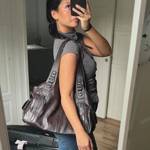 Mörkbrun axelväska i skinnlook - Snygg mörkbrun handväska med axelremmar och flera dragkedjefickor. Väskan har en mjuk, slouchy form och detaljer i silverfärgad metall. Perfekt för dig som vill ha en rymlig och stilren väska till vardags. Materialet är konstläder.