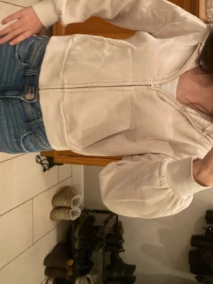 Beige hoodie med dragkedja - Säljer en beige hoodie med dragkedja och stora fickor framtill. Hoodien har en klassisk huva och ribbade muddar vid ärmar och nederkant. Perfekt för en avslappnad och chill stil. Materialet känns mjukt och bekvämt, troligen bomull eller bomullsblandning.