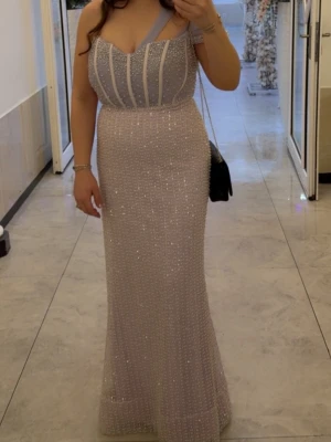 Glittrig långklänning med pärldetaljer - Elegant långklänning i ljus lillafärg med glittriga pärldetaljer och smala axelband. Klänningen har figurnära passform och markerad midja, samt en v-ringad urringning och dekorativa vertikala linjer upptill. Perfekt för speciella tillfällen.