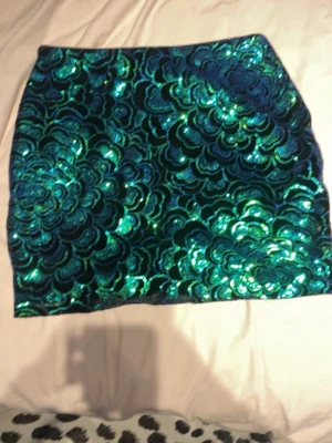 Grönblå glittrig paljettkjol - Säljer en kort kjol täckt av gröna och blå paljetter i ett blommigt mönster. Kjolen har en tajt passform och är perfekt för dig som vill sticka ut med en färgstark och glittrig look. Materialet är syntet och paljetterna ger ett riktigt festligt intryck.