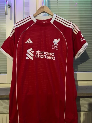 Liverpool FC matchtröja Adidas röd - Säljer en röd Liverpool FC matchtröja från Adidas med vita detaljer och broderad LFC-logga på bröstet. Tröjan har vita ränder på axlarna, sponsortryck på framsidan och klassisk sportig passform. Perfekt för dig som älskar fotboll och vill stötta ditt lag.
