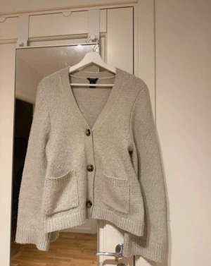 Beige stickad kofta med fickor - Mysig beige stickad kofta med v-ringning och tre stora knappar framtill. Koftan har två praktiska fickor och ribbade muddar vid ärmslut och nederkant. Perfekt för lager på lager och enkel att matcha med olika outfits.