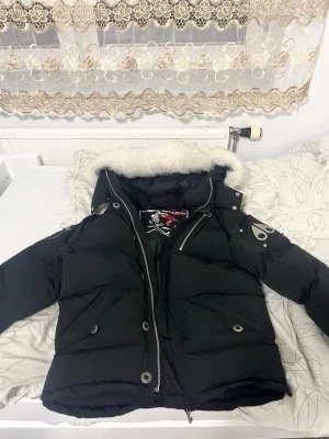 Äkta Svart dunjacka Moose Knuckles - Snygg svart dunjacka från Moose Knuckles med avtagbar huva och vit fuskpäls. Jackan har dragkedja, tryckknappar och två fickor framtill. Ikonisk logga på ärmen och coolt innerfoder med tryck. Perfekt för kalla dagar. 