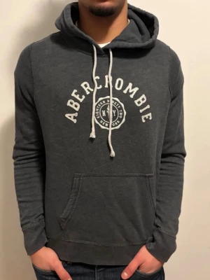 Sällsynt Mörkgrå hoodie från Abercrombie & Fitch - Snygg mörkgrå hoodie från Abercrombie med vit logga och text på bröstet. Klassisk känguruficka framtill, justerbar huva med vita snören och mjukt material som känns skönt mot huden. Perfekt för en avslappnad och trendig look. EXTREMT EFTERTRAKTAD MODELL
