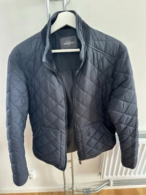 Marinblå quiltad jacka från Dressmann - Snygg svart quiltad jacka från Dressmann med dragkedja framtill och ribbad krage. Jackan har ett diskret rutmönster och är tillverkad i polyester, perfekt för kyligare dagar. Ena innerfickan har gått sönder, se bild 