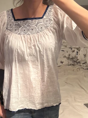 Vit blus med blå brodyr från Vila - Söt vit blus från Vila med blå broderade detaljer runt den fyrkantiga halsringningen och blå kantband. Blusen har lång ärm och är gjord i ett tunt, luftigt material som ger en avslappnad look. Perfekt att styla med jeans eller kjol.