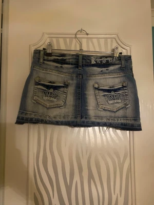 Kort jeanskjol från K&B Denim - Säljer en kort jeanskjol från K&B Denim med slitna detaljer, stora fickor både fram och bak samt dragkedjor framtill. Kjolen har en klassisk blå tvätt och broderade loggor på bakfickorna. Perfekt för dig som gillar streetstyle och denim. Tror storlek S-M
