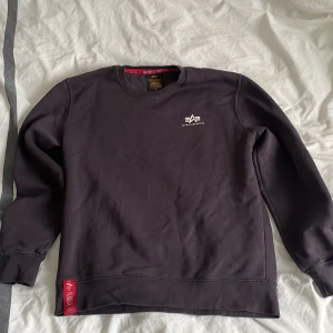  sweatshirt från Alpha Industries - Säljer en svart sweatshirt från Alpha Industries i storlek M. Tröjan har rund hals, diskret logga på bröstet och en röd detalj vid nederkanten. Tillverkad i mjuk bomullsmix med borstad insida för extra komfort. Perfekt för en clean och stilren look.