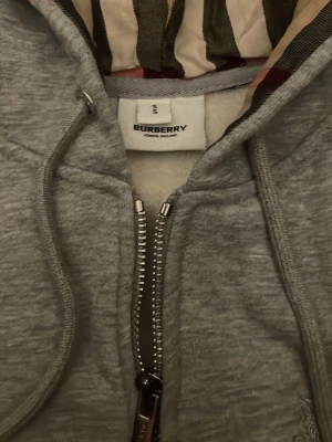 Grå zip hoodie från Burberry - Snygg grå hoodie från Burberry med dragkedja framtill och klassiskt rutigt foder i huvan. Tillverkad i mjukt bomullsmaterial och har justerbar dragsko i huvan. Perfekt för en avslappnad och stilren look.