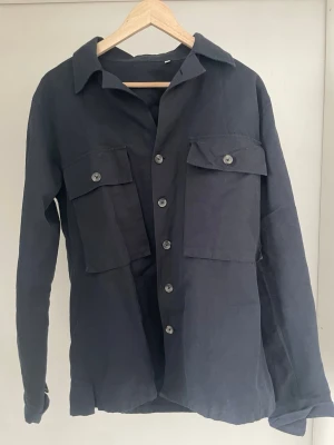 Mörkblå overshirt från Albini 1876 - Stilren mörkblå overshirt från Albini 1876 med knappar framtill och två stora bröstfickor med lock. Skjortkrage och långa ärmar med knapp vid ärmslut. Tillverkad i ett mjukt, vävt material som ger en avslappnad look. Perfekt att bära öppen eller stängd över en t-shirt.