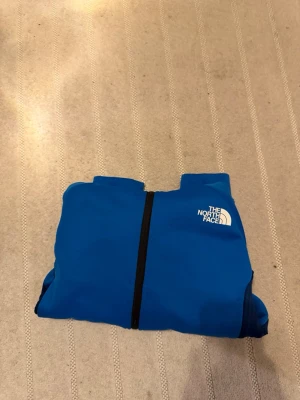 Blå vindjacka från The North Face - Snygg blå vindjacka med huva från The North Face. Jackan har svart dragkedja, vita logotyper på bröstet och ärmen samt paneler i mörkare blått på sidorna. Perfekt för blåsiga dagar och enkel att packa ner i väskan.
