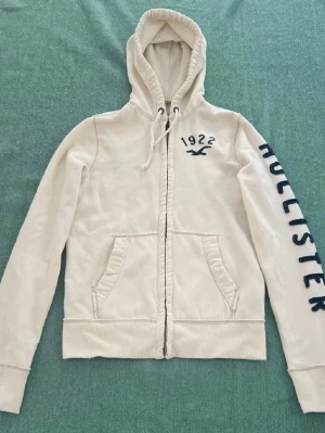 Vit hoodie från Hollister med tryck - Snygg vit (HERR) hoodie från Holliste med dragkedja, huva med dragsko och två fickor fram. Mörkblått tryck och logga på bröstet samt stort HOLLISTER-tryck längs ena ärmen.