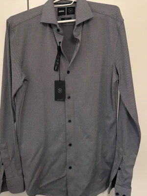 Slim fit skjorta från BOSS - Snygg slim fit skjorta från BOSS i gråvitt småmönstrat tyg. Skjortan har klassisk krage, långa ärmar och svarta knappar. Perfekt för dig som gillar stilrena och moderna plagg. Passar M och L