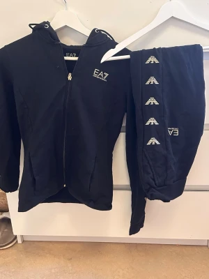 Svart EA7 Emporio Armani set - Snyggt svart set från EA7 Emporio Armani med zip hoodie och matchande byxor. Setet har vita loggor på både tröjan och byxbenet, samt huva på tröjan. Perfekt för en sportig och avslappnad stil. Materialet känns mjukt och bekvämt.