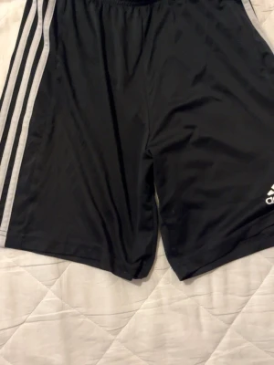 Svarta Adidas träningsshorts - Svarta träningsshorts från Adidas med klassiska vita ränder längs sidorna och Adidas-logga på ena benet. Tillverkade i ett lätt och snabbtorkande material som passar perfekt för sport och träning.