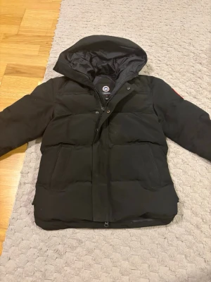 Svart dunjacka från Canada Goose - Snygg svart dunjacka från Canada Goose med huva och klassisk logotyp på ärmen. Jackan har två stora framfickor, dragkedja och tryckknappar framtill. Perfekt för kalla vinterdagar och riktigt varm tack vare dunfyllningen.