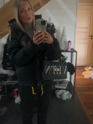 Svart pufferjacka från Fjällräven - Säljer en svart pufferjacka från Fjällräven med klassisk logga på bröstet och gula detaljer på dragkedjorna. Jackan har hög krage och stora fickor framtill, perfekt för kalla vinterdagar. Snygg och trendig modell som håller dig varm.