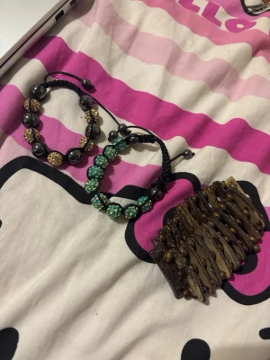 Set med tre färgglada armband - Säljer ett set med tre armband: ett svart med stora pärlor och glittriga guldfärgade kulor, ett grönt med skimrande pärlor och ett brett armband i brun plast med träliknande struktur. Alla har justerbara band och cool design som sticker ut. Allad e tre kostar 20 kr och för en kostar det bara 10 kr