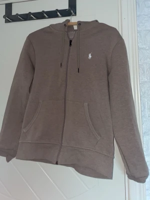 Beige ziphoodie från Ralph Lauren - Säljer en beige hoodie från Ralph Lauren med dragkedja framtill och två fickor. Klassisk vit logga broderad på bröstet och justerbar huva med snören. Tillverkad i mjukt bomullsmaterial, perfekt för en avslappnad och stilren look. Använt knappast mer än 6 gånger