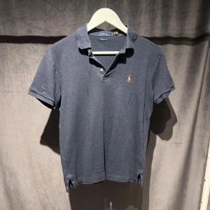 Mörkblå pikétröja från Ralph Lauren - Klassisk mörkblå pikétröja från Ralph Lauren med korta ärmar och den ikoniska broderade loggan på bröstet. Tröjan har krage, knappar vid halsen och är tillverkad i mjuk bomull. Perfekt till både jeans och shorts.