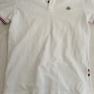 Vit Moncler pikétröja med logga - Snygg vit pikétröja från Moncler med klassisk krage och knappar framtill. Tröjan har Moncler-logga på bröstet och detaljer i rött och blått på ärmsluten. Perfekt för en stilren och sportig look.