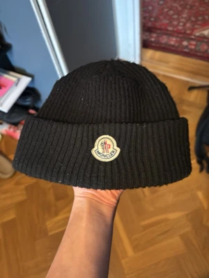 Svart ribbstickad mössa från Moncler - Säljer en svart ribbstickad mössa från Moncler med uppvikt kant och Moncler-logga framtill. Klassisk och stilren design som passar perfekt till kalla dagar. Mössan är gjord i ett mjukt och värmande material.