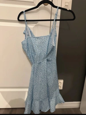 Blå blommig omlottklänning - Söt blå kortklänning med små vita blommor och volangdetaljer. Klänningen har omlottmodell, smala axelband och en lätt, luftig känsla – perfekt för varma dagar. Volanger längs kanten ger en extra feminin touch.