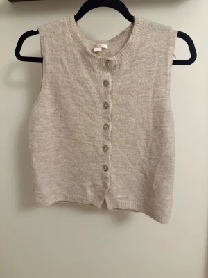 Beige stickad väst med knappar - Mysig beige stickad väst med rund hals och sju knappar framtill. Perfekt att styla över en skjorta eller t-shirt för en trendig lager-på-lager-look. Enkel och stilren design som passar till mycket.
