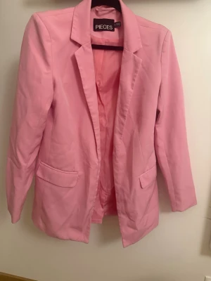 Rosa kavaj från PIECES - Snygg rosa kavaj från PIECES med klassisk krage och två fickor framtill. Kavajen har en rak passform och är helfodrad för extra komfort. Perfekt för dig som vill addera färg till din outfit.