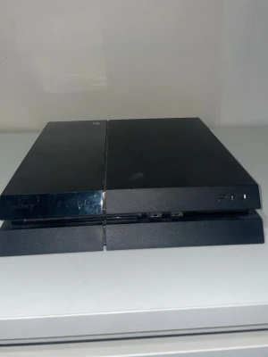 Sony PlayStation 4 spelkonsol svart - Säljer en svart PlayStation 4 från Sony. Konsolen har en stilren, rektangulär design med matta och blanka ytor. På framsidan finns USB-portar och på baksidan HDMI, nätverksport och strömingång. Perfekt för gaming och streaming. PlayStation 4 kontroll ingår