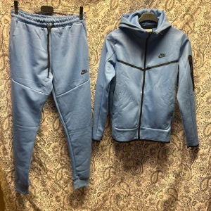 Nike blått tracksuit set - Mysigt tracksuit set från Nike i ljusblått. Setet består av en hoodie med dragkedja och svarta detaljer samt matchande byxor med resår i midjan och smal passform. Båda delarna har Nike-logga och är tillverkade i mjukt, fleeceliknande material.