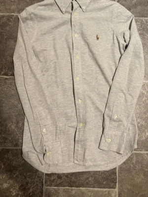 Grå skjorta från Ralph Lauren - Klassisk grå skjorta från Ralph Lauren med broderad logga på bröstet. Skjortan har lång ärm, knappar framtill och button-down krage. Perfekt för en clean och stilren look.