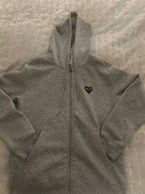 Comme des Garçons Zip Hoodie - Snygg grå zip-hoodie från Comme des Garçons. Har ett svart hjärta på fram o baksidan av tröjan. Säljer pga att den inte kommer till användning längre. Fått i Julklapp, köpt på NK Stockholm. Använt cirka 5 gånger. Ställ så många frågor ni vill! Fraktar samma dag. Priset är ej hugget i sten!💯📦🚛