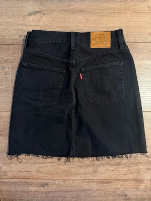 Svart jeanskjol från Levi's - Snygg svart jeanskjol från Levi's med rå kant nertill och klassiska fem fickor. Kjolen har bälteshällor, knapp- och dragkedjestängning samt detaljer i kopparfärg. Perfekt för en avslappnad och trendig look.