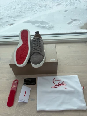 Brun/grå sneakers från Christian Louboutin - Snygga gråa sneakers i mocka från Christian Louboutin med klassisk röd sula och vita kanter. Skorna har gråa skosnören och en stilren design som passar till det mesta. Kommer med originalkartong, dustbag och skohorn.