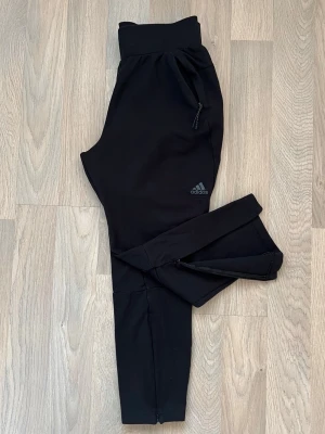 Träningsbyxor från Adidas med dragkedja - Svarta träningsbyxor från Adidas med dragkedjor vid bensluten och fickorna för extra prestanda. Byxorna har en bred resår i midjan och atletisk passform. Diskret Adidas-logga på vänster lår.  Byxorna är tillverkade i ett mjukt och stretchigt material vilket gör dem perfekta för träning (t.ex fotboll, gym) eller bara att ha hemma. Använda bara fåtal gånger. Hör gärna av dig vid frågor.