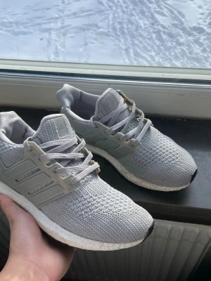 Grå Adidas Ultraboost sneakers - Snygga grå Adidas Ultraboost sneakers med stickad ovandel i mesh och klassiska tre ränder på sidan. Skorna har vita Boost-sulor och grå snören. Perfekta för dig som gillar sportig och clean stil. Skorna kommer utan box.