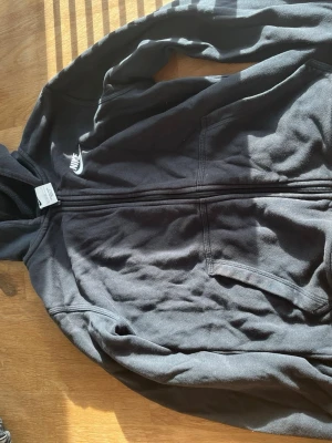 Svart zip hoodie från Nike - Säljer en svart hoodie från Nike med dragkedja framtill och vit broderad logga på bröstet. Hoodien har två fickor fram och en klassisk huva. Tillverkad i mjukt bomullsmaterial som är skönt att ha på sig. Ordinarie pris är 500 