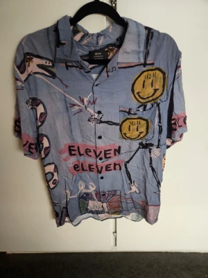 Blå kortärmad skjorta från Neuw - Unik blå skjorta från Neuw med färgglatt abstrakt mönster och smileys. Skjortan har korta ärmar, klassisk krage och knappar framtill. Perfekt för dig som vill sticka ut med en kreativ och lekfull stil.