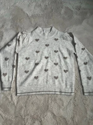 Grå stickad tröja med hjärtan - Supersöt ljusgrå stickad tröja från Freequent med små ljusbruna hjärtan över hela. Tröjan har puffade axlar, ribbade muddar och rund halsringning. Perfekt för dig som gillar gulliga detaljer och vill ha något mysigt till hösten eller vintern. Använd 1 gång, pris kan diskuteras 