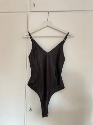 Svart body med smala axelbanb - Snygg svart body i glansigt material med smala justerbara axelband och djup urringning fram och bak. Perfekt att styla till olika outfits för en cool vibe.