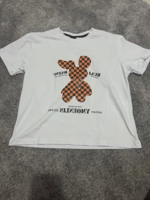 Vit t-shirt med schackrutig kanin - Vit t-shirt med tryck av en orange och svart schackrutig kanin på framsidan. T-shirten har korta ärmar och rund halsringning. Trycket har även textdetaljer. Perfekt för en cool och avslappnad stil.