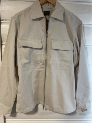 Beige overshirt från Jack & Jones - Stilren beige overshirt från Jack & Jones Premium med grov struktur, dragkedja framtill och två stora bröstfickor med lock. Klassisk krage och knapp vid ärmslut. Perfekt lager på lager-plagg i ett robust bomullsmaterial.