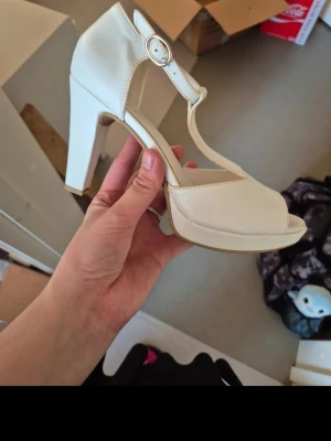 Vita pumps med klack från Catwalk - Snygga vita pumps från Catwalk med hög klack och öppen tå. Skon har ett justerbart spänne runt ankeln och en stadig plattform framtill. Tillverkad i syntetmaterial med stilren design som ger en elegant look.