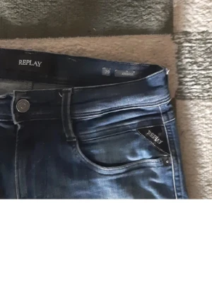 Blå Replay Anbass jeans, stl 29/32 - Snygga blå Replay Anbass jeans med klassisk femficksdesign och diskreta slitningar. Jeansen har normal passform och är tillverkade i mjukt bomullsmaterial med lite stretch för extra komfort. Perfekta för dig som gillar en stilren och modern look.
