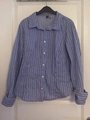 Blåvit randig skjorta med knappar - Klassisk blå skjorta med vita och svarta vertikala ränder. Skjortan har lång ärm, krage och knäppning framtill med vita knappar. Tillverkad i bomull för en skön och luftig känsla. Perfekt till jeans eller chinos för en clean look.